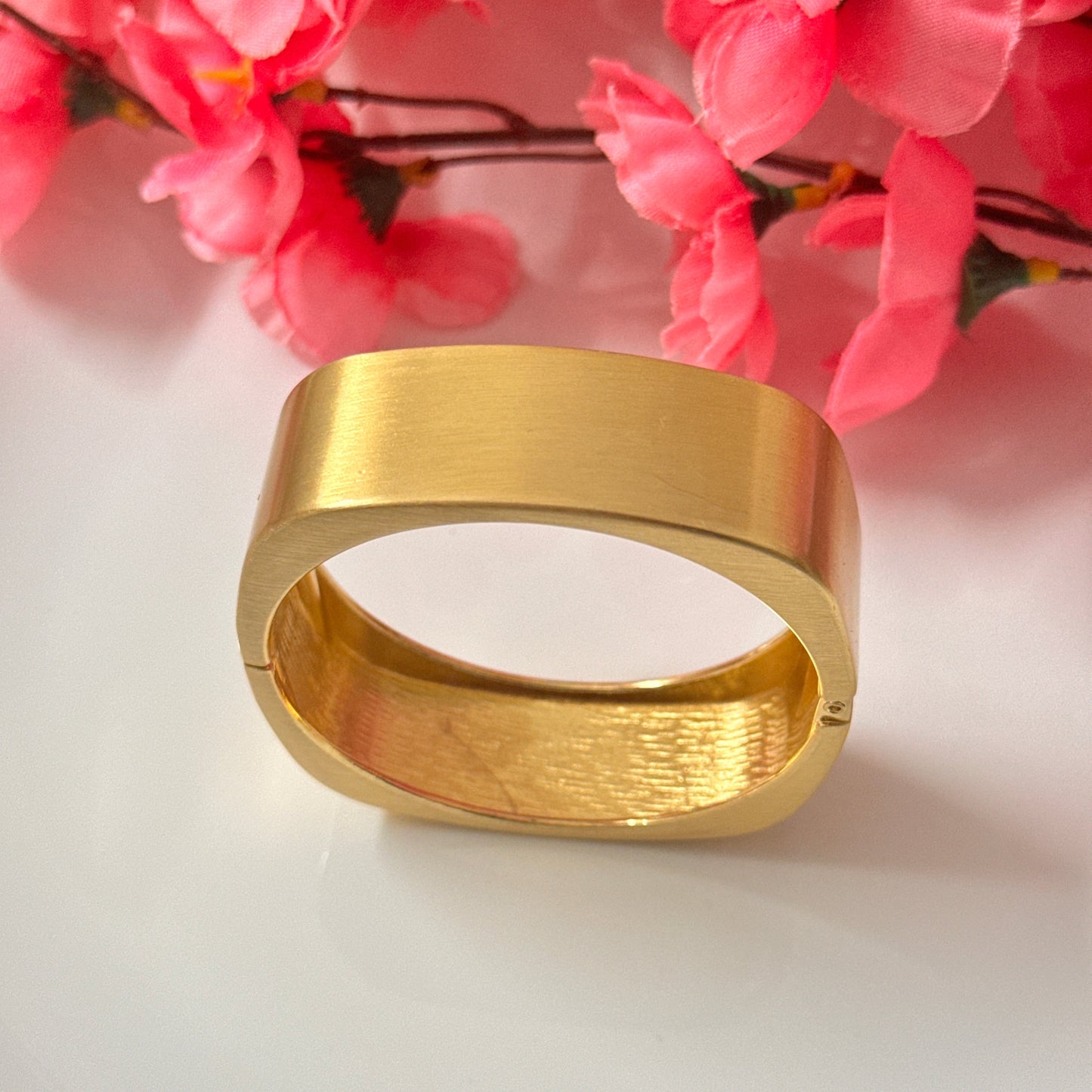 A gold chunky bracelet displayed on a white surface with pink floral decoration in the background.