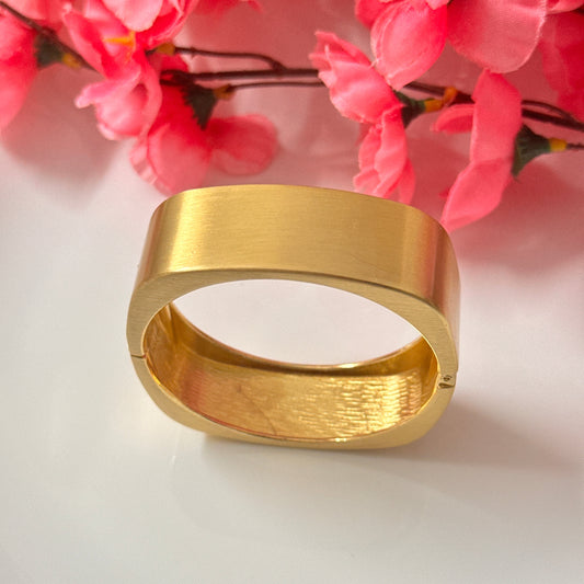A gold chunky bracelet displayed on a white surface with pink floral decoration in the background.