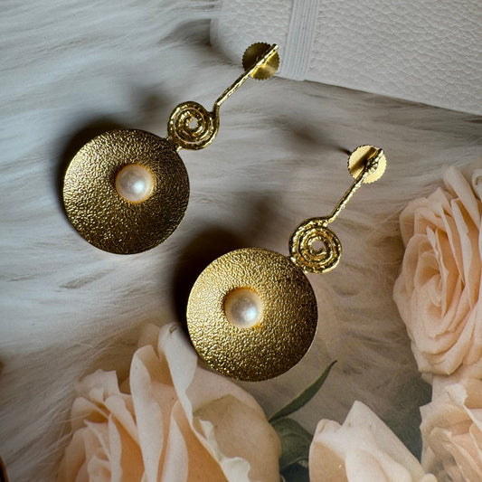 A pair of golden brass pearl stud earrings displayed on a faux fur surface with pink flowers in the background.