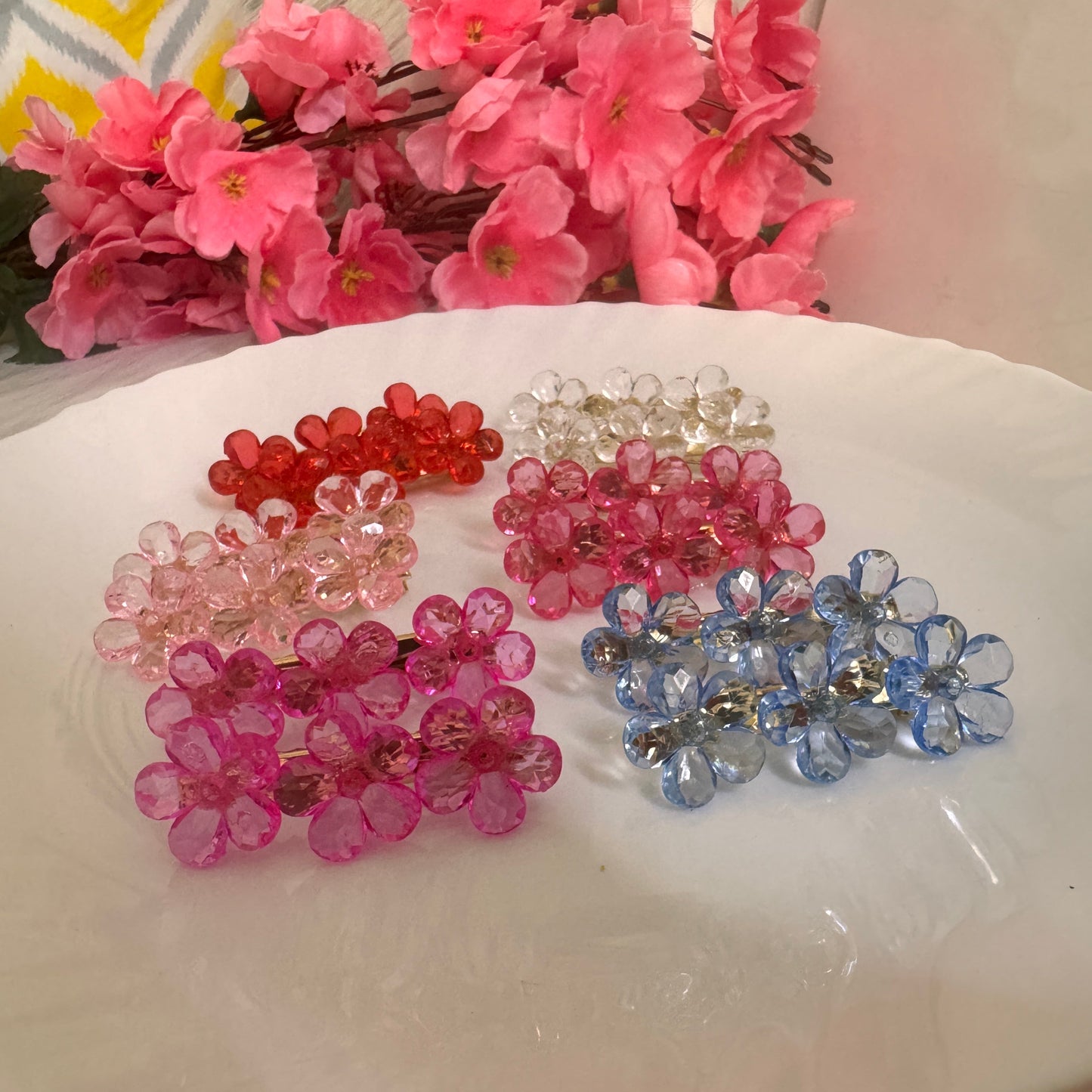 A set of crystal floral clips in various colors displayed on a white surface, with pink flowers in the background.