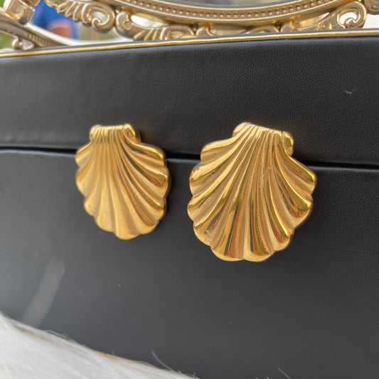 A pair of golden shell-shaped stud earrings placed on a black surface, with a reflective surface above them.