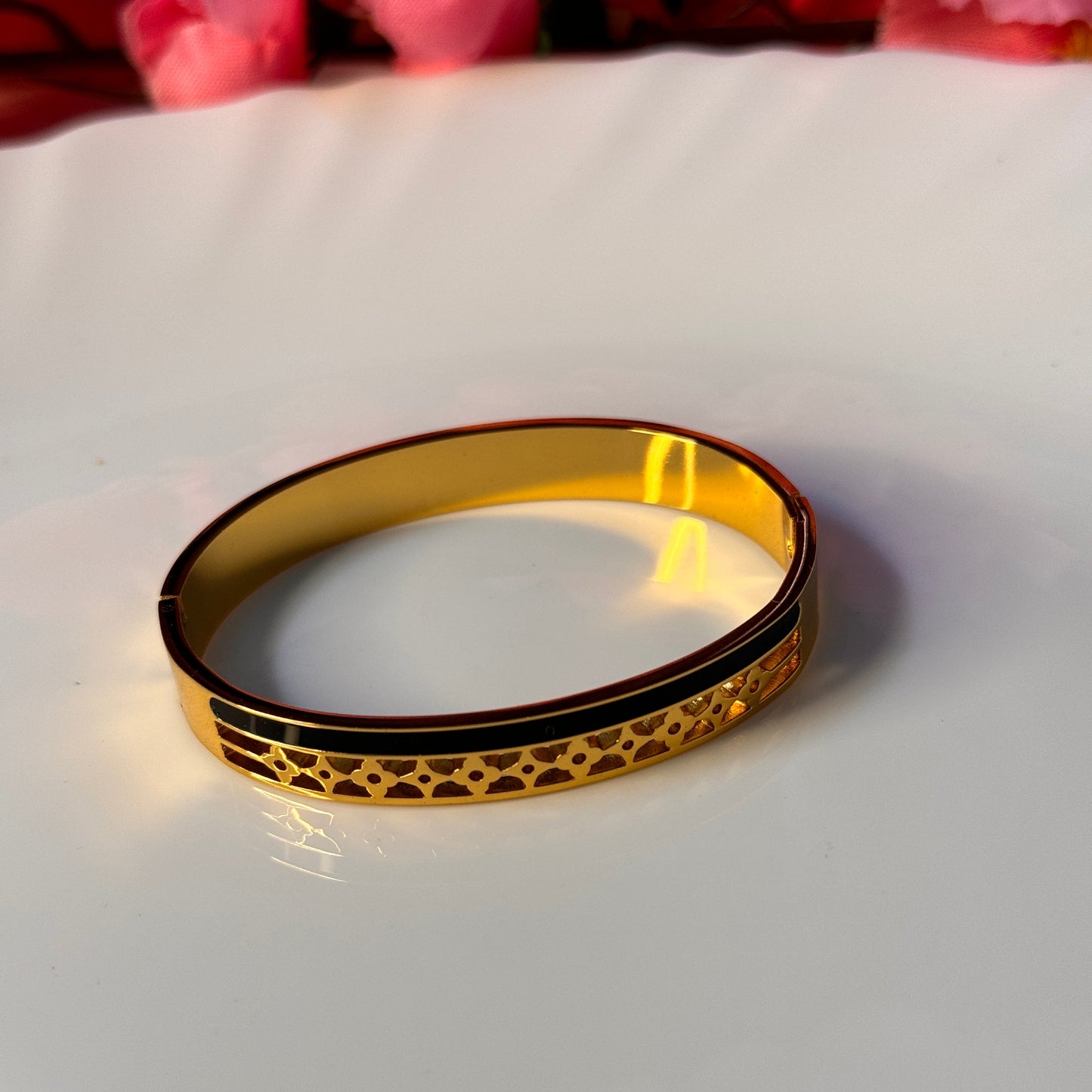 A golden bracelet with a patterned design, displayed on a white surface with red flowers in the background.
