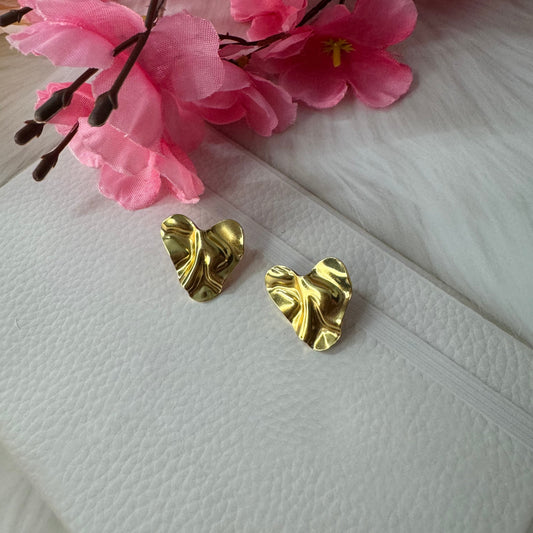 A pair of golden chunky heart-shaped stud earrings displayed on a white surface with pink floral decor in the background.