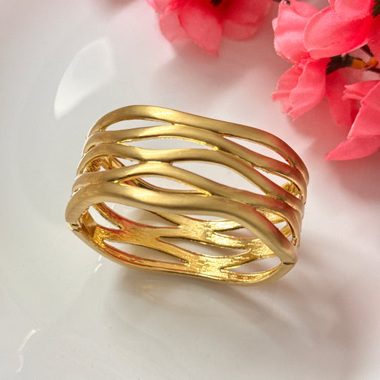 A gold-plated cuff bracelet with a cutout pattern, displayed on a white surface with pink flowers in the background.