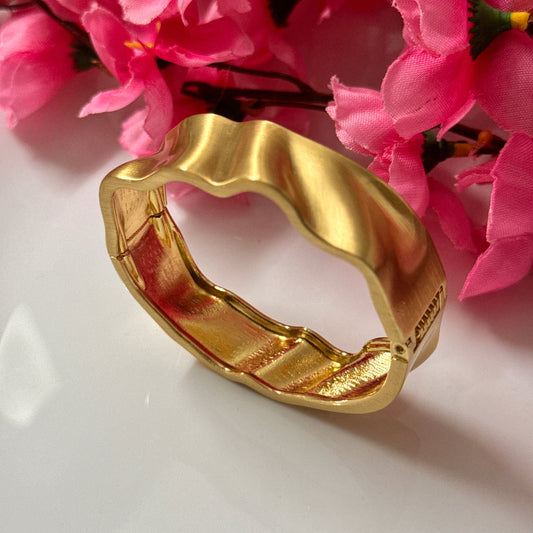 A gold-plated bangle bracelet with a matte finish, displayed alongside pink floral decor.