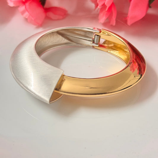 A gold and silver tone bracelet displayed on a white surface with pink flowers in the background.
