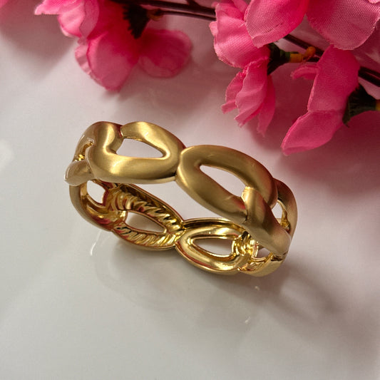 A gold-colored chunky cuff bracelet with a frosted metallic shine, displayed against a white background with pink flowers.