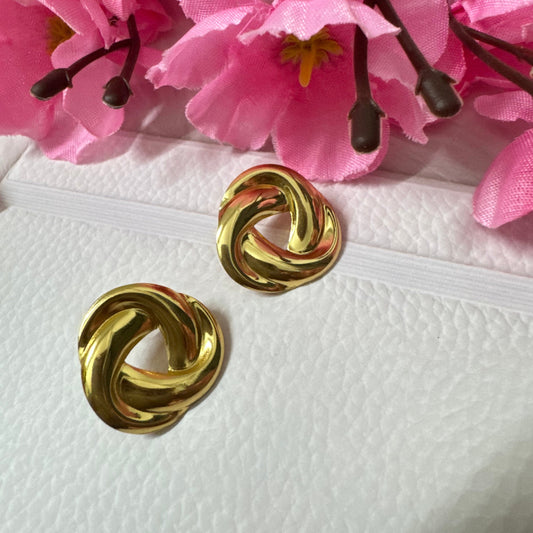 A pair of gold-colored knotted designer studs placed on a white surface with pink floral background.