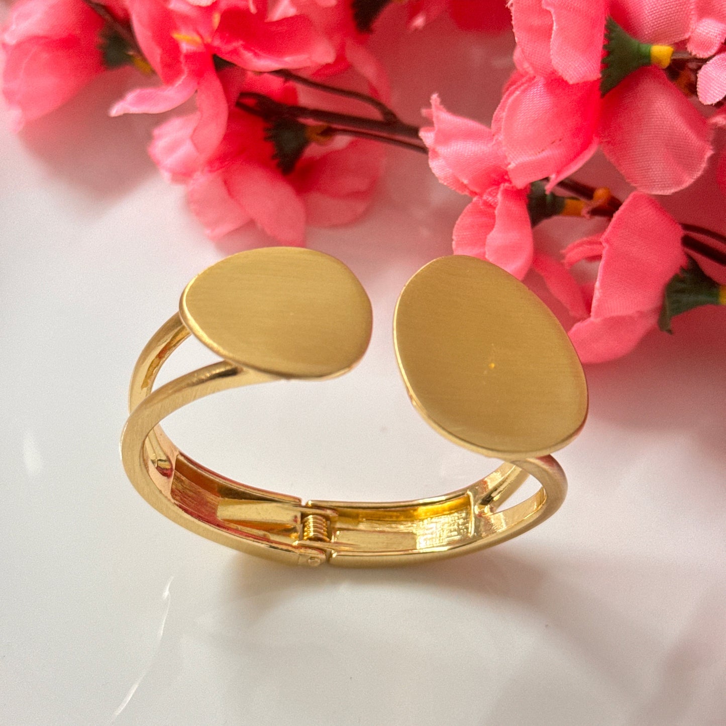 A golden chunky bracelet displayed on a white surface with pink flowers in the background.