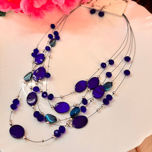 A layered necklace with blue and green beads displayed on a white plate with pink flowers in the background.