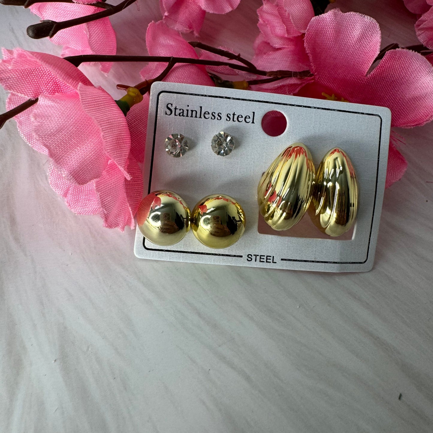 A set of three golden hoop earrings on a white card with 'Stainless steel' text, displayed alongside pink floral decor.