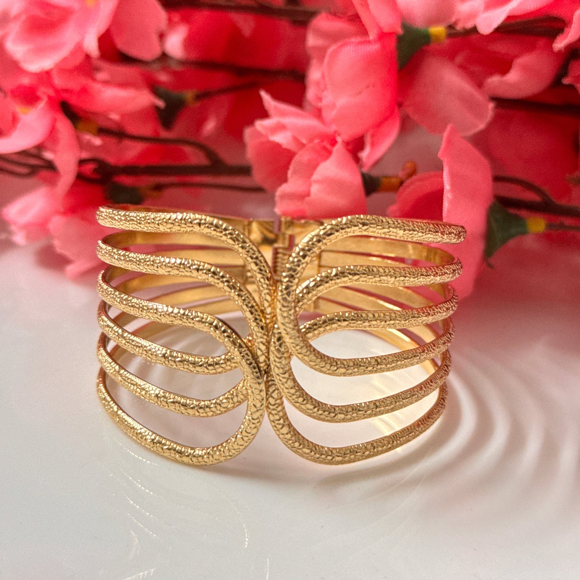 A gold-colored hand cuff with a textured surface, displayed against a backdrop of pink flowers.