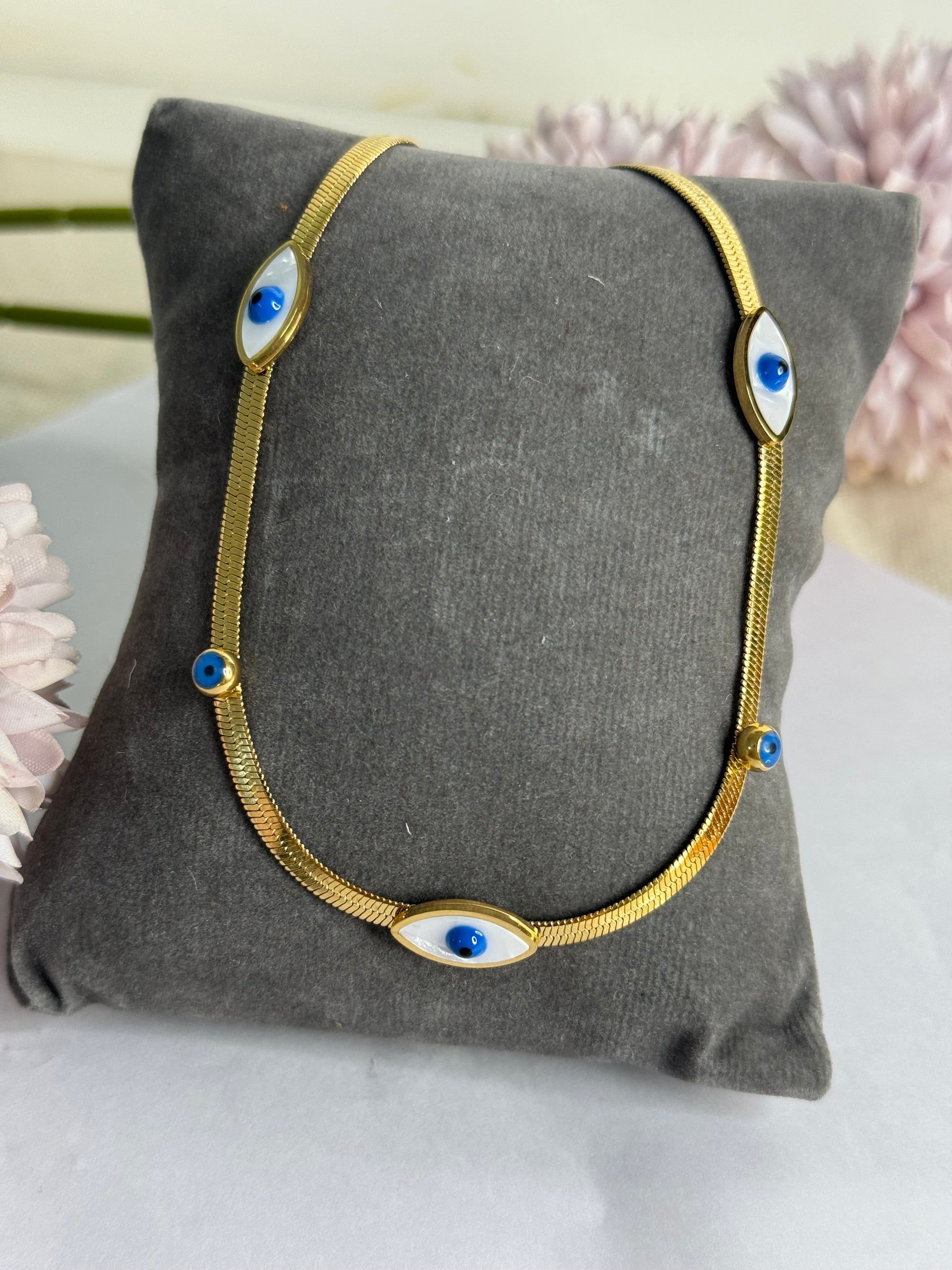 A gold-plated chain necklace with a blue and white evil eye design displayed on a gray cushion.