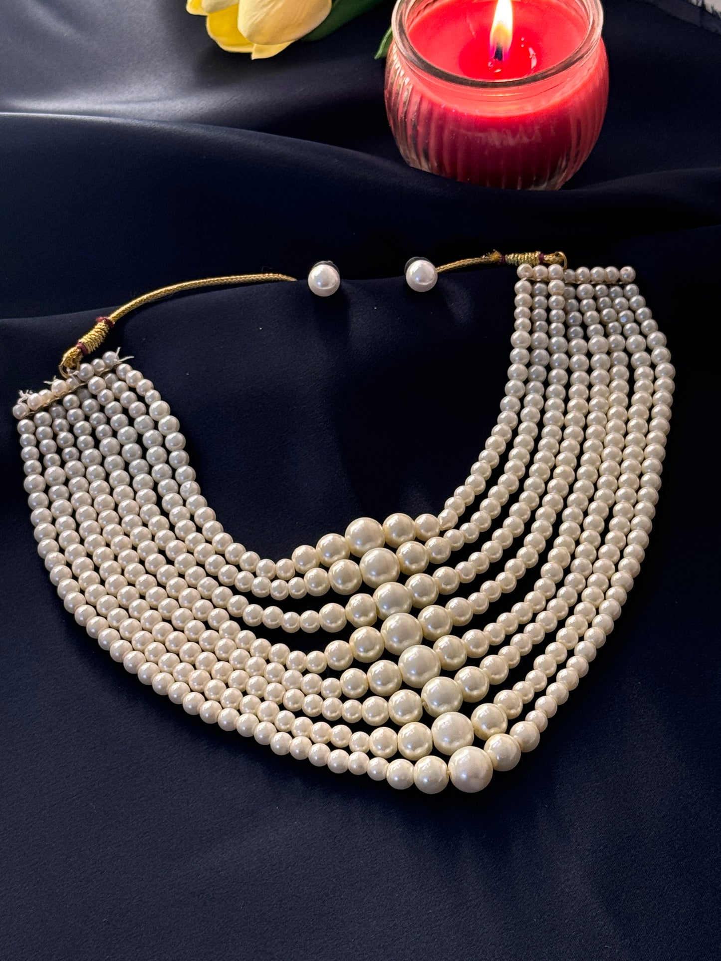 White beaded necklace on a dark fabric background with a red candle and yellow flower.