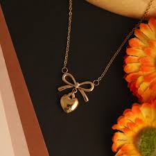 Gold necklace with a heart and bow pendant on a dark background with orange flowers.