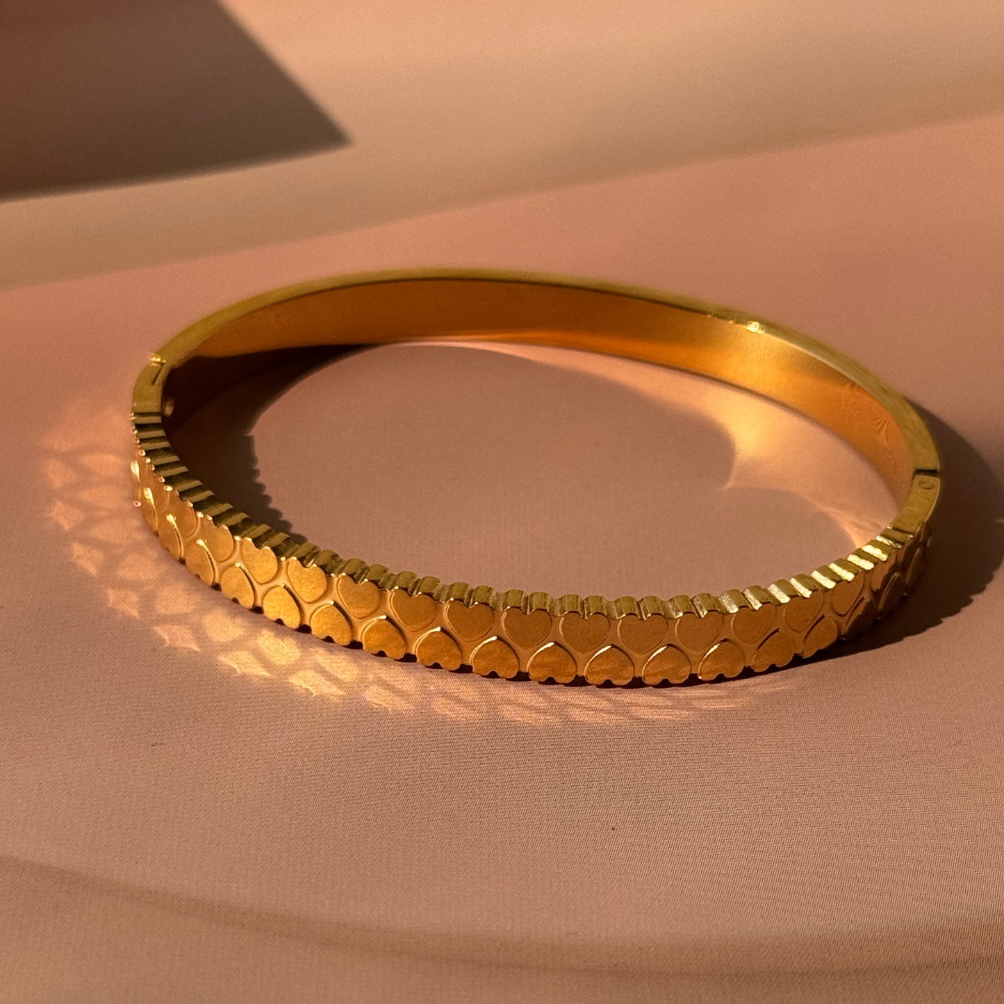 A gold-colored bracelet with heart-shaped studs, displayed against a beige background.