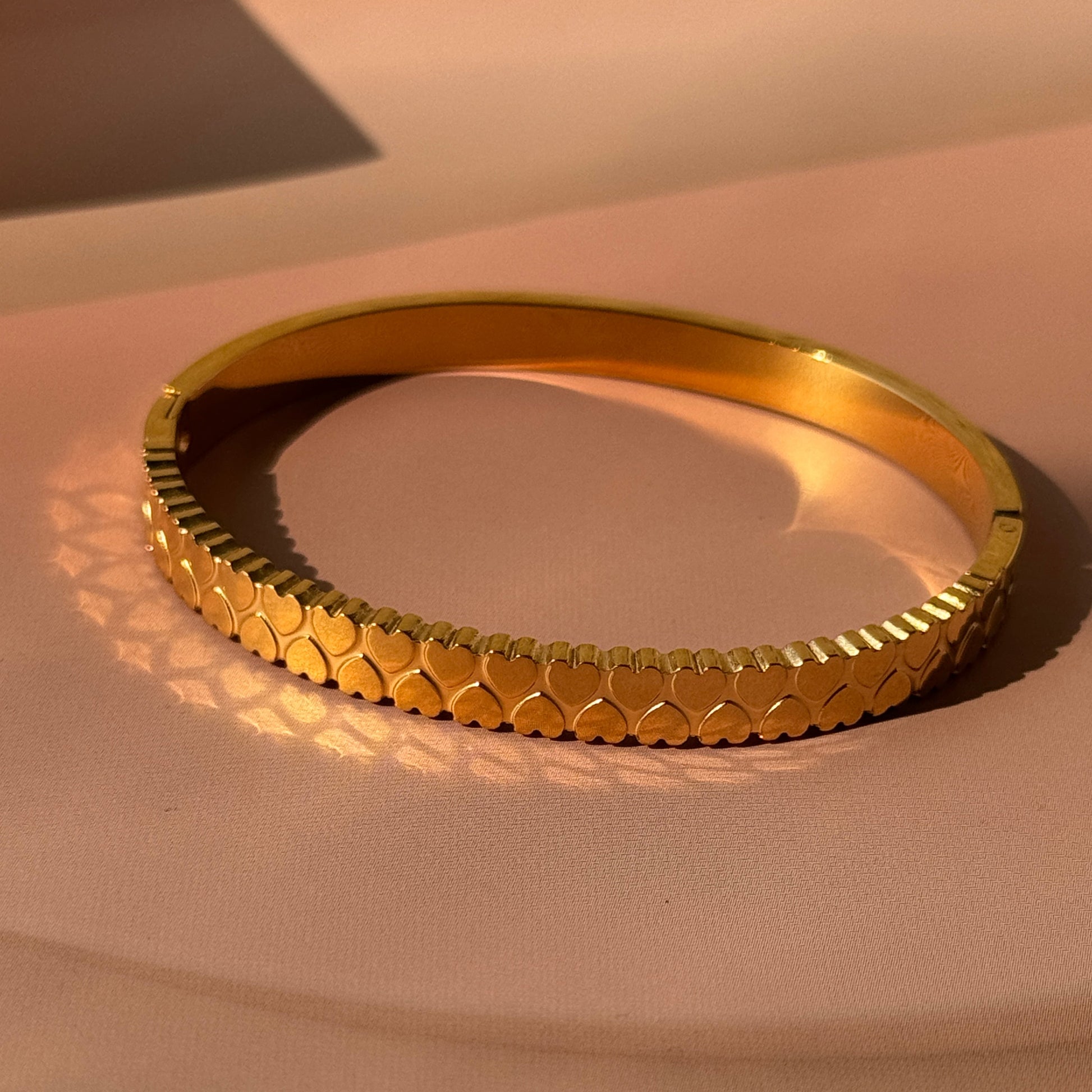 A gold-colored bracelet with heart-shaped studs, displayed against a beige background.