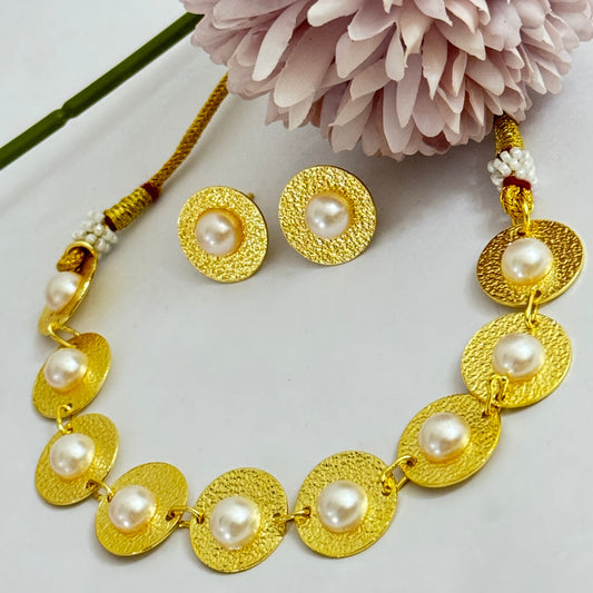 A gold-colored necklace with coin-shaped pearl beads and floral accents, displayed on a white surface next to a pink flower.