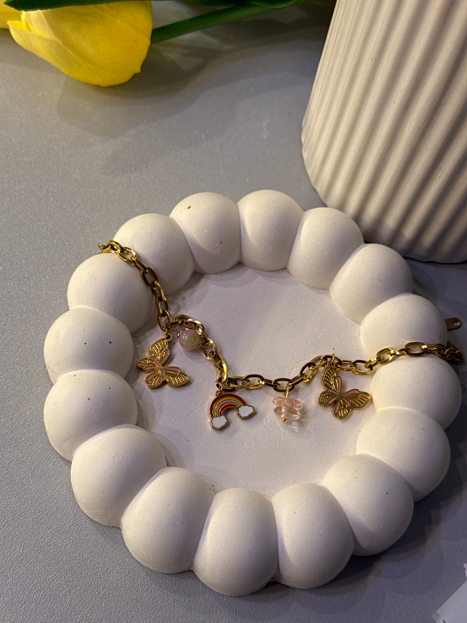 White beaded bracelet with gold charms on a white dish, yellow flower in the corner