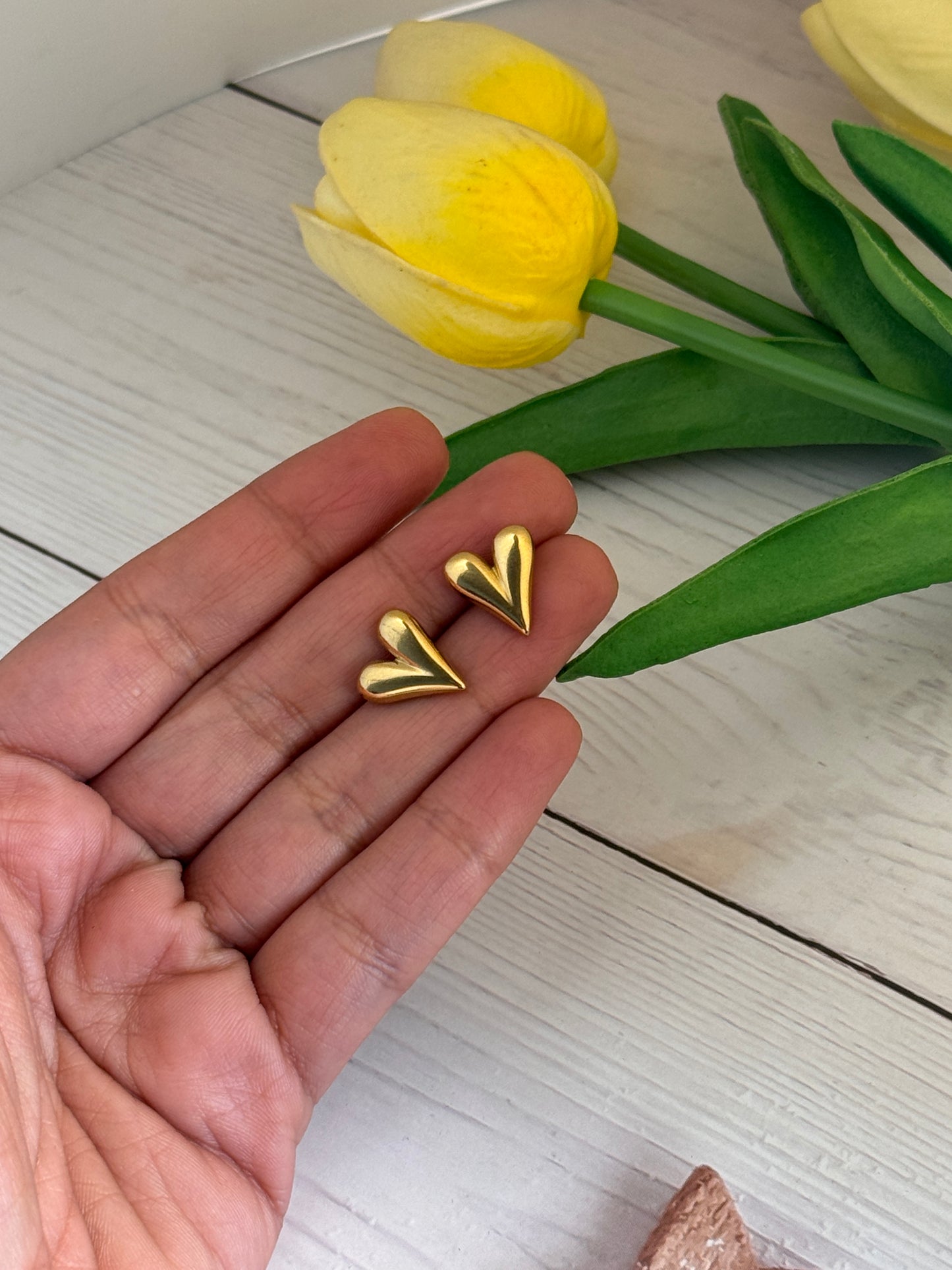 Gold heart-shaped earrings held in a hand with yellow tulips on a light wooden surface