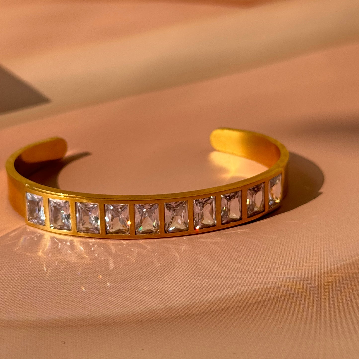 A gold-colored bracelet with clear square stones set in a row, displayed against a light background.