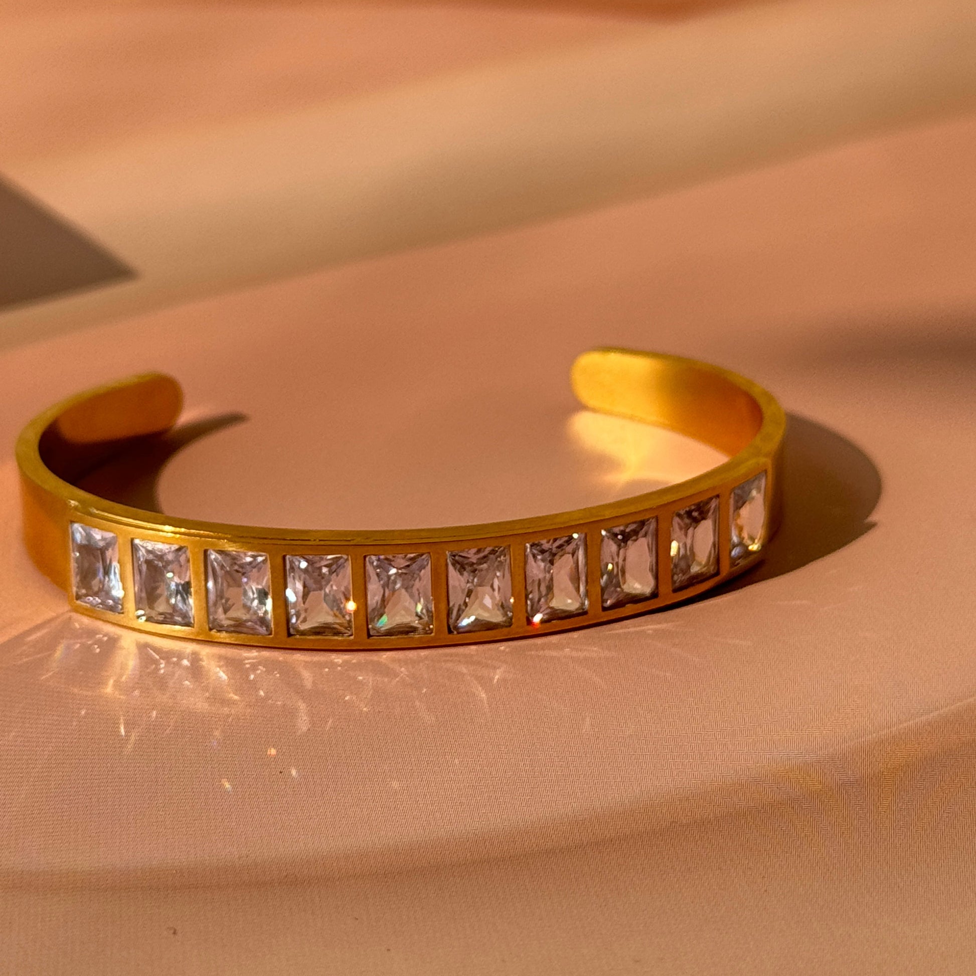 A gold-colored bracelet with clear square stones set in a row, displayed against a light background.