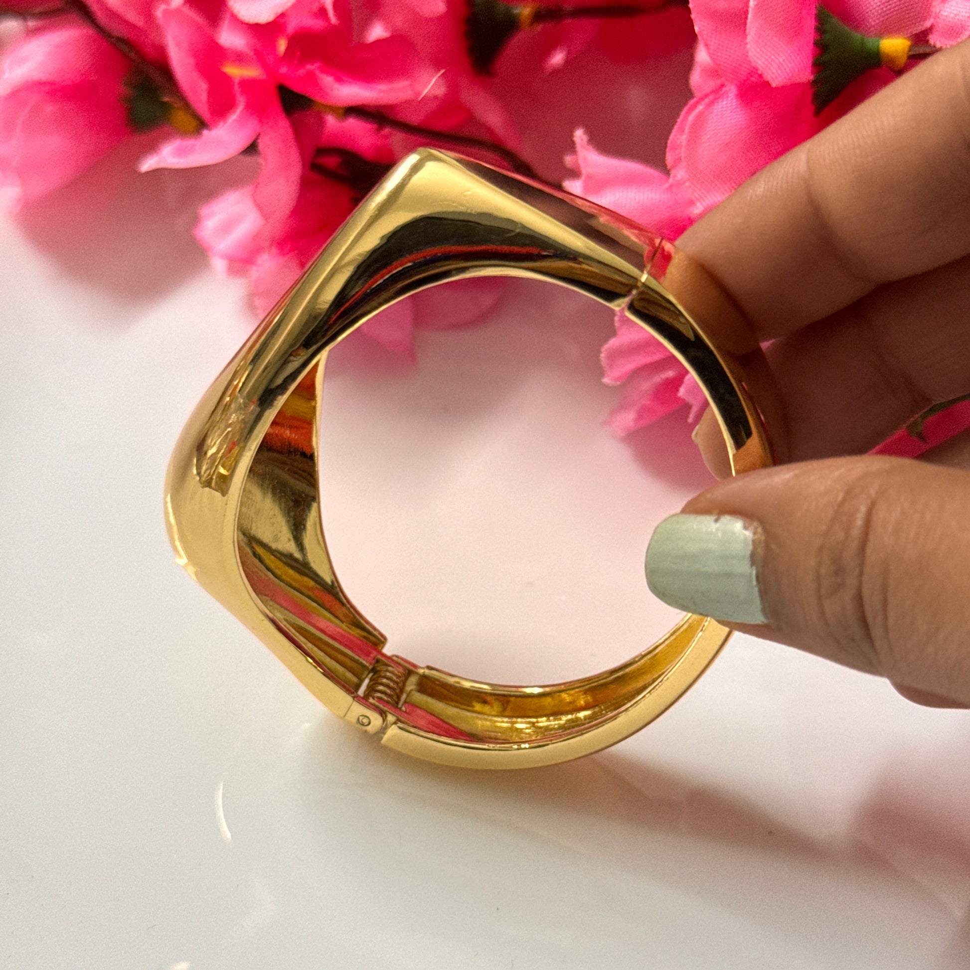A gold-colored tear drop open bracelet being held by a person with pink flowers in the background.