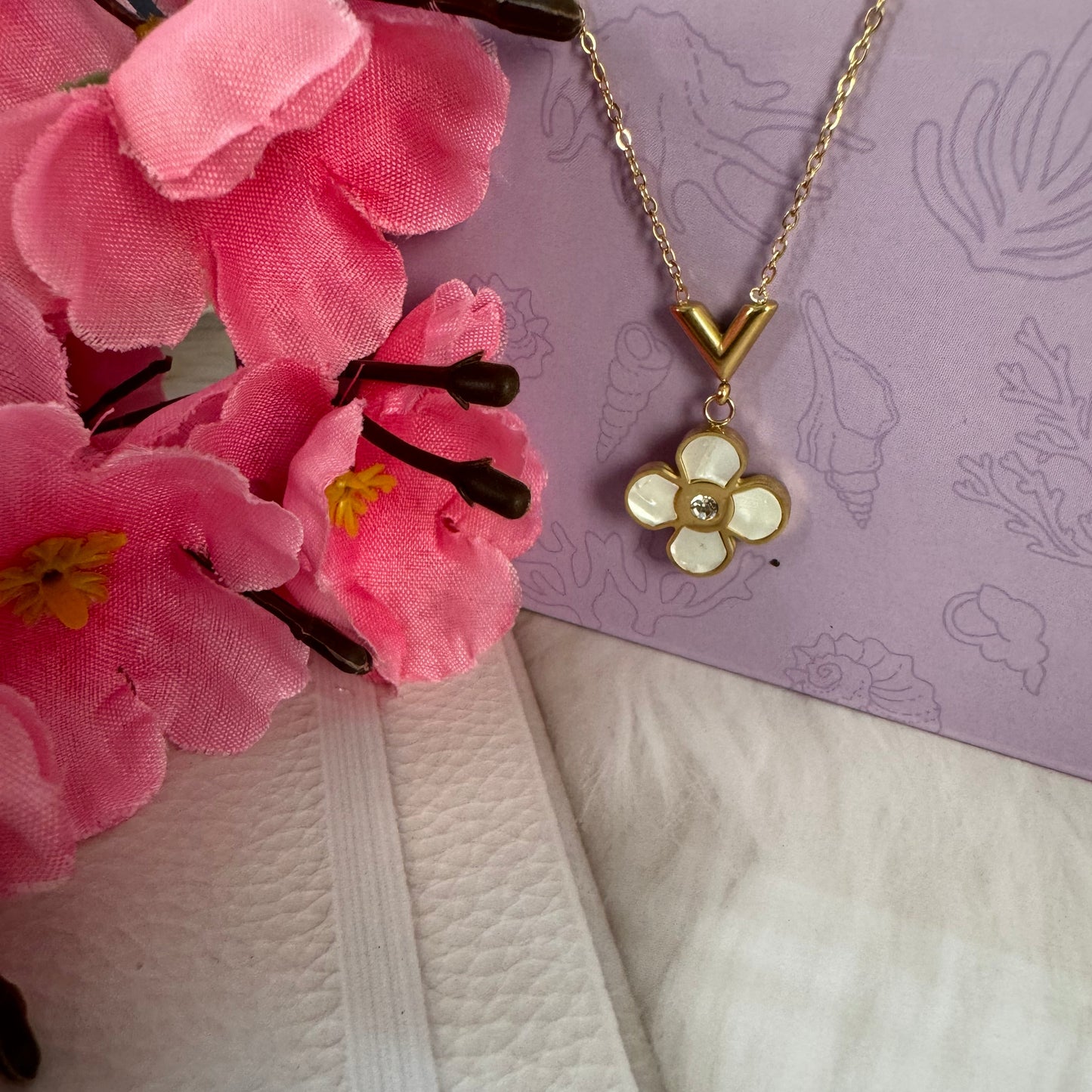 A gold-colored necklace with a floral pendant displayed on a purple background with a pink floral pattern and a white element to the side.