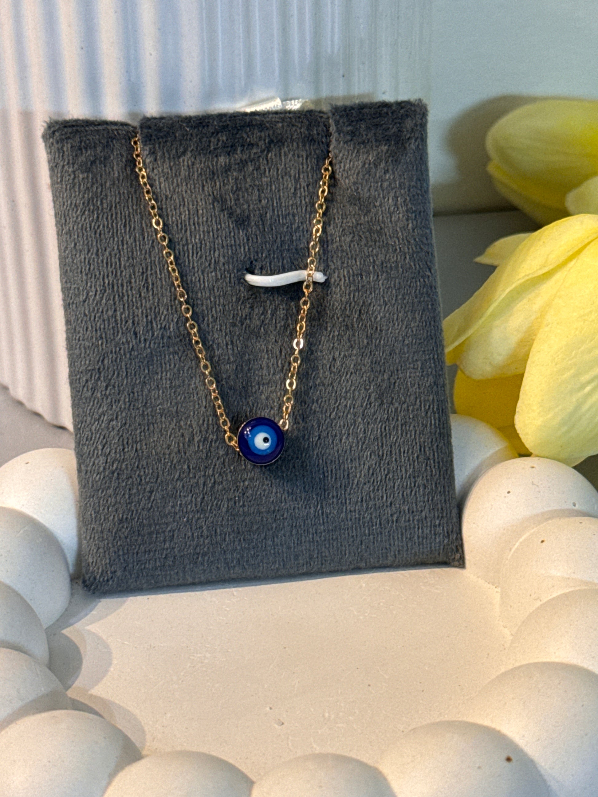 Necklace with a blue evil eye charm on a gray card with yellow flowers in the background