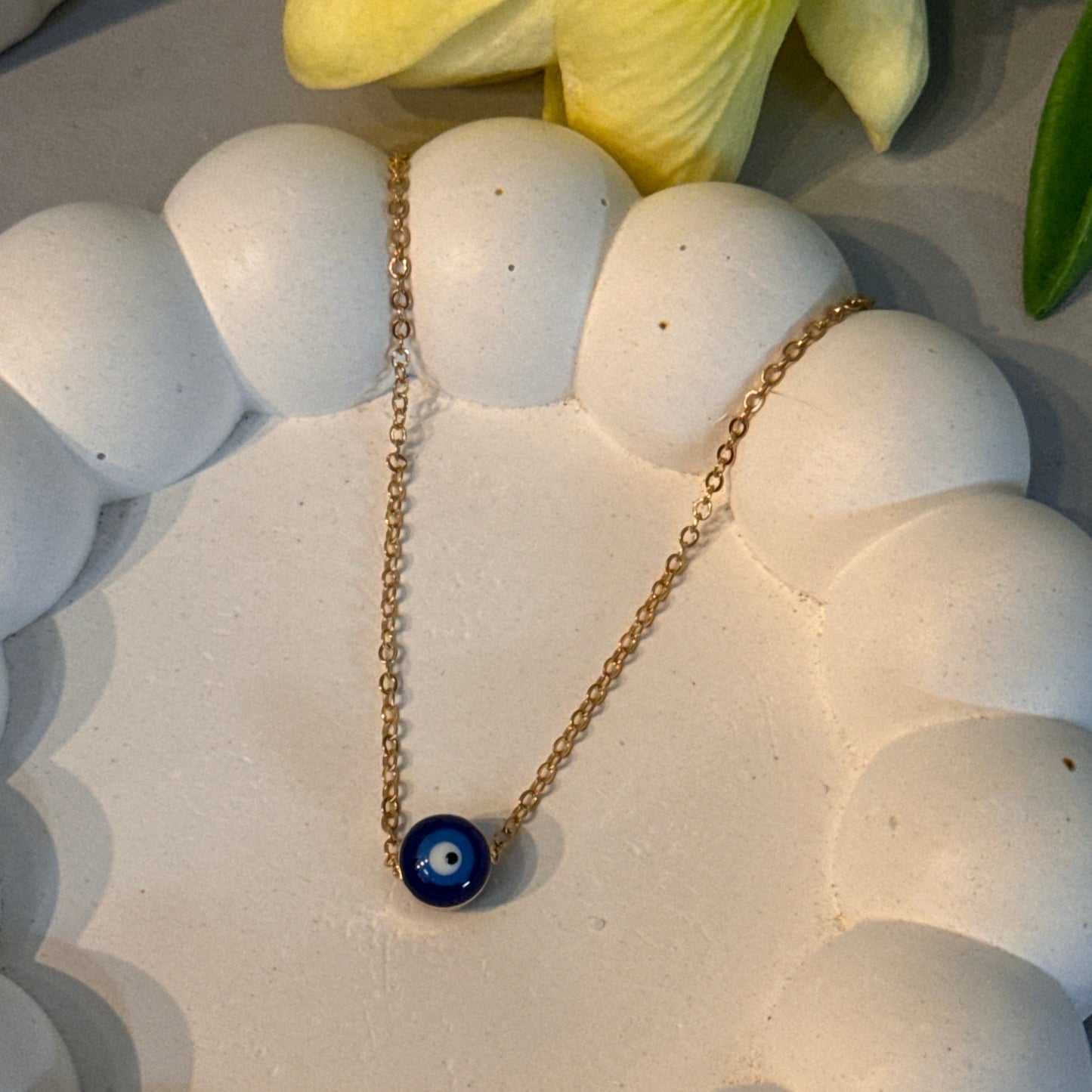 Gold necklace with a blue pendant on a white stone surface next to yellow flowers.
