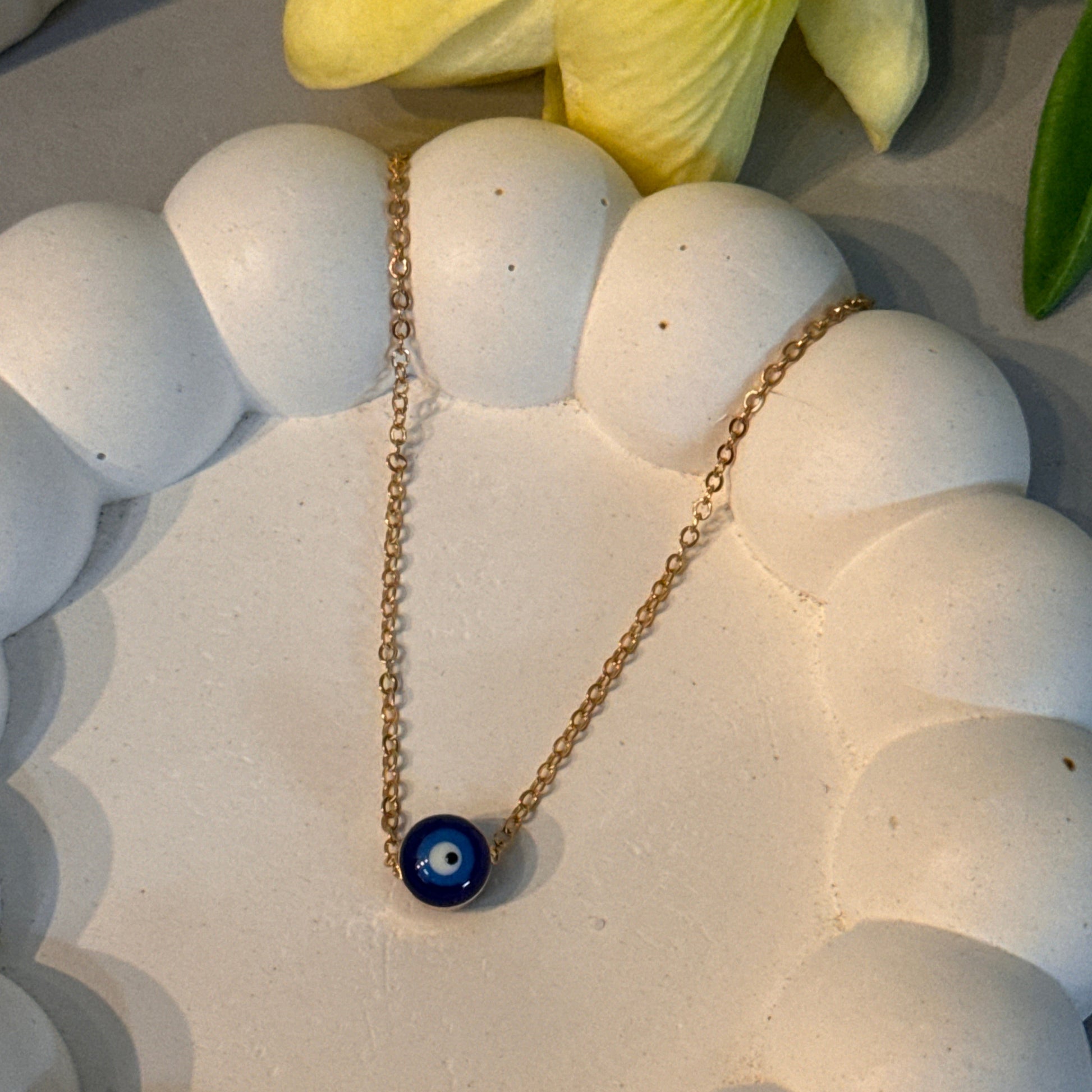 Gold necklace with a blue pendant on a white stone surface next to yellow flowers.