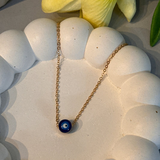 Gold necklace with a blue pendant on a white stone surface next to yellow flowers.
