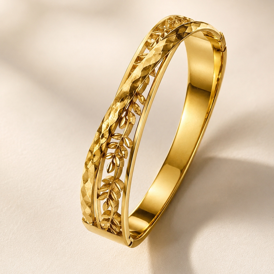 A polished gold bangle bracelet with a textured, woven leaf-like pattern on one side, displayed on a soft beige background with elegant shadows.