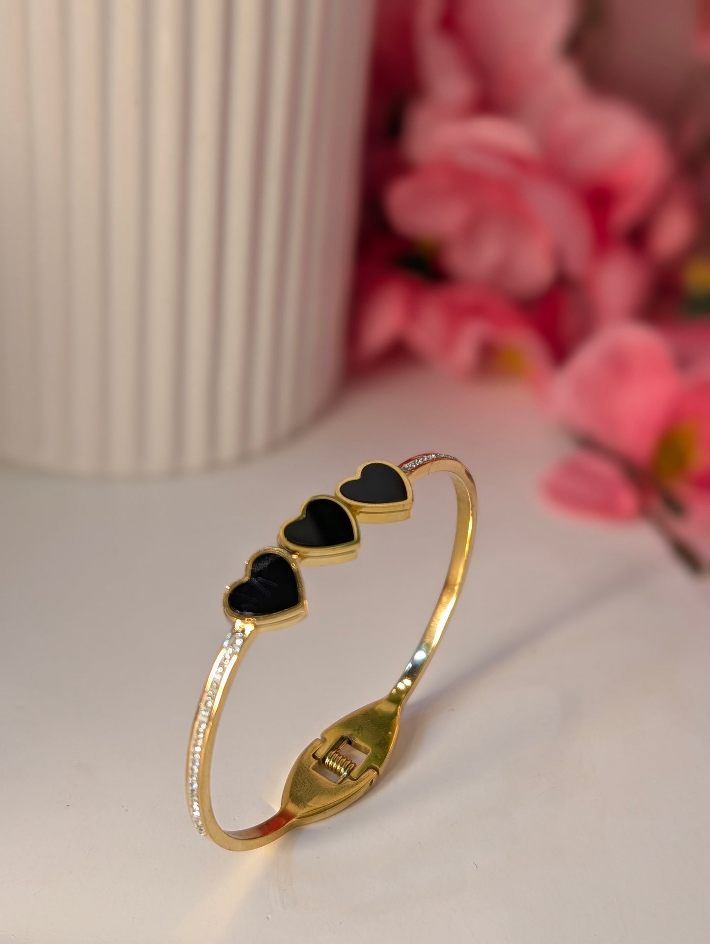 Gold bracelet with black heart charms on a light surface with pink flowers in the background