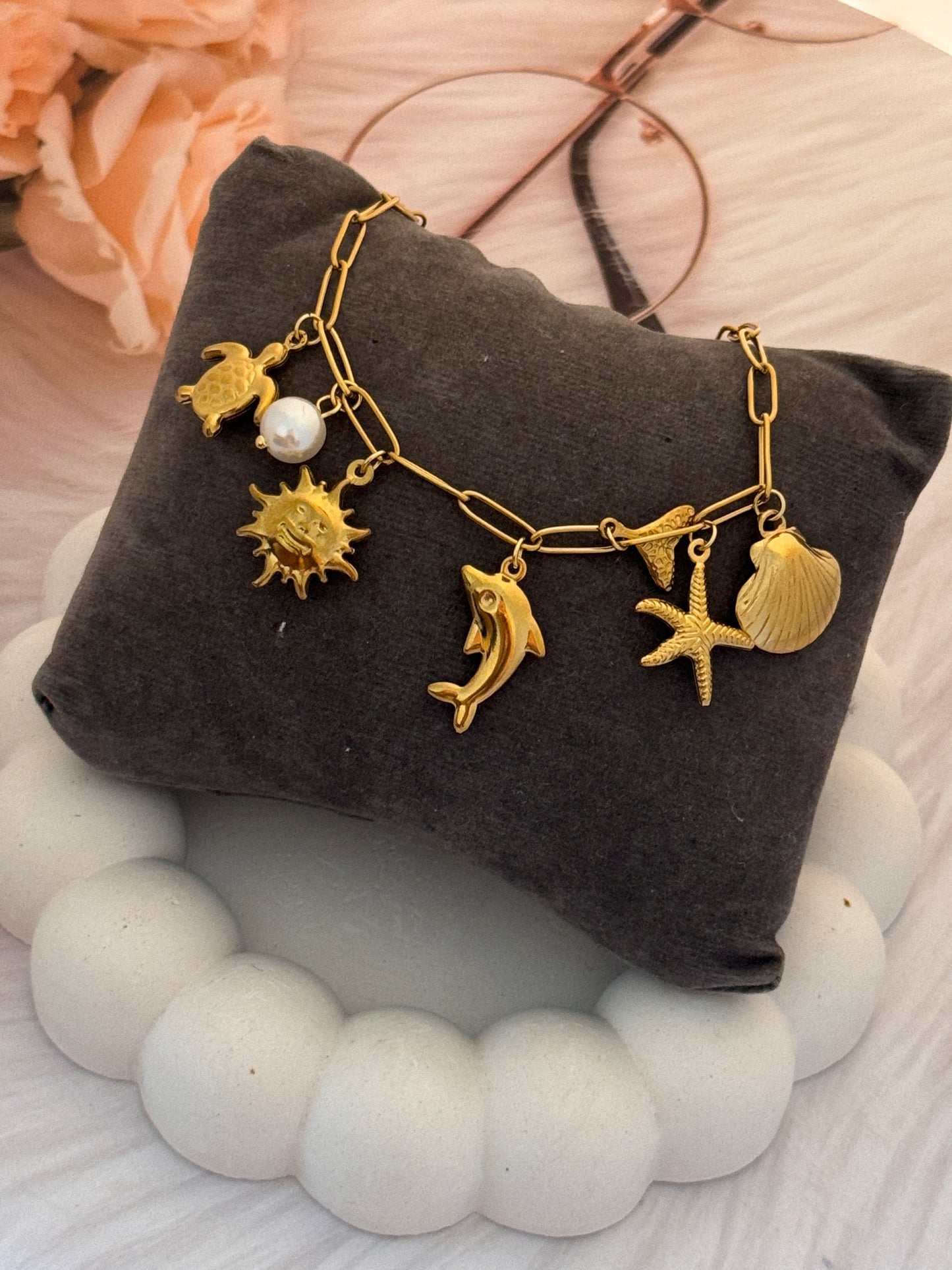 Gold charm bracelet on a dark fabric pillow with decorative stones and flowers.