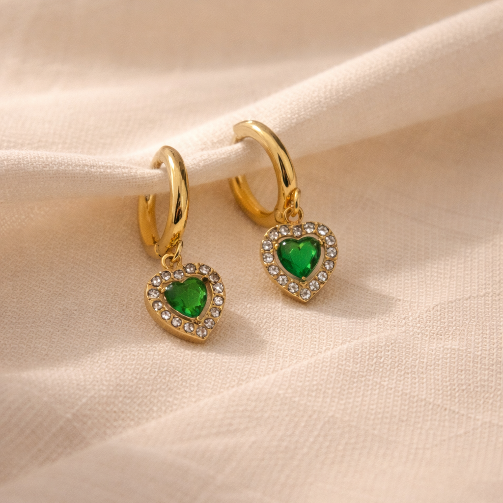 Gold heart-shaped earrings with green gemstones on a beige fabric background