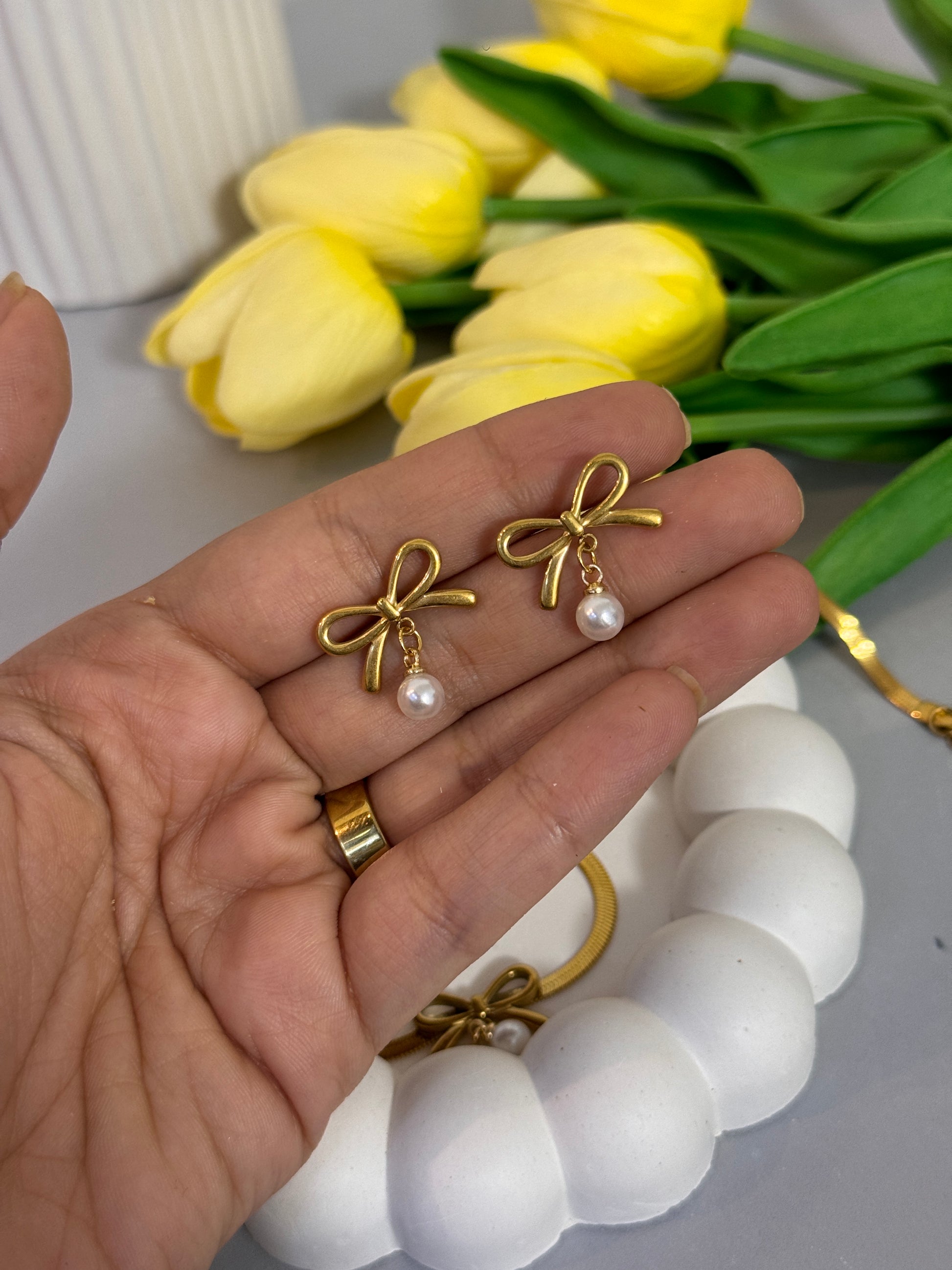 Gold bow-shaped earrings with pearl centers held in a hand, with yellow tulips and a white bracelet in the background.