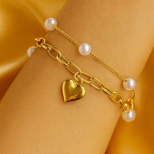 Gold chain bracelet with heart charm and pearls on a wrist against a yellow fabric background