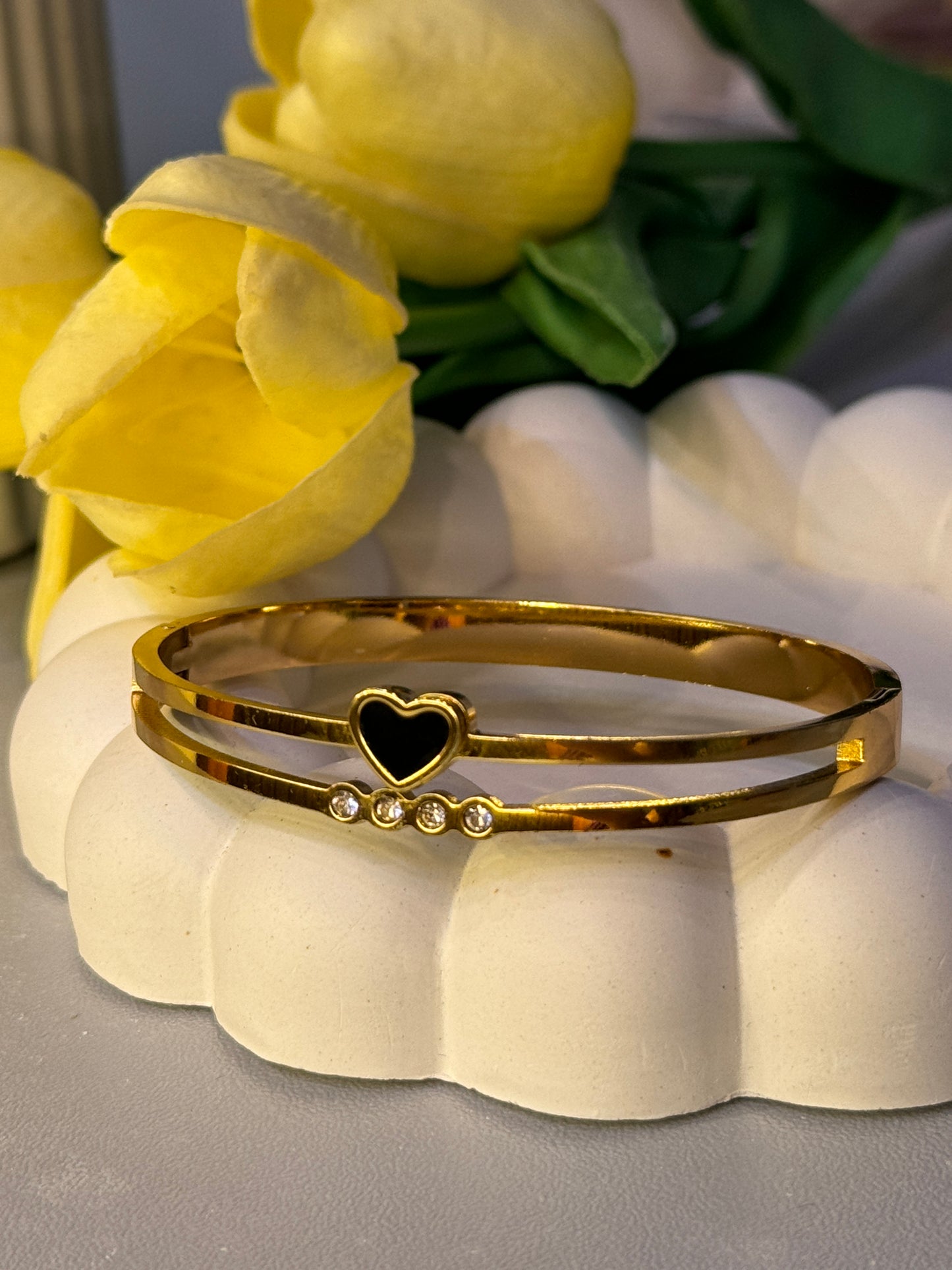 Gold bracelet with heart and black stone design on a white stand with yellow flowers in the background