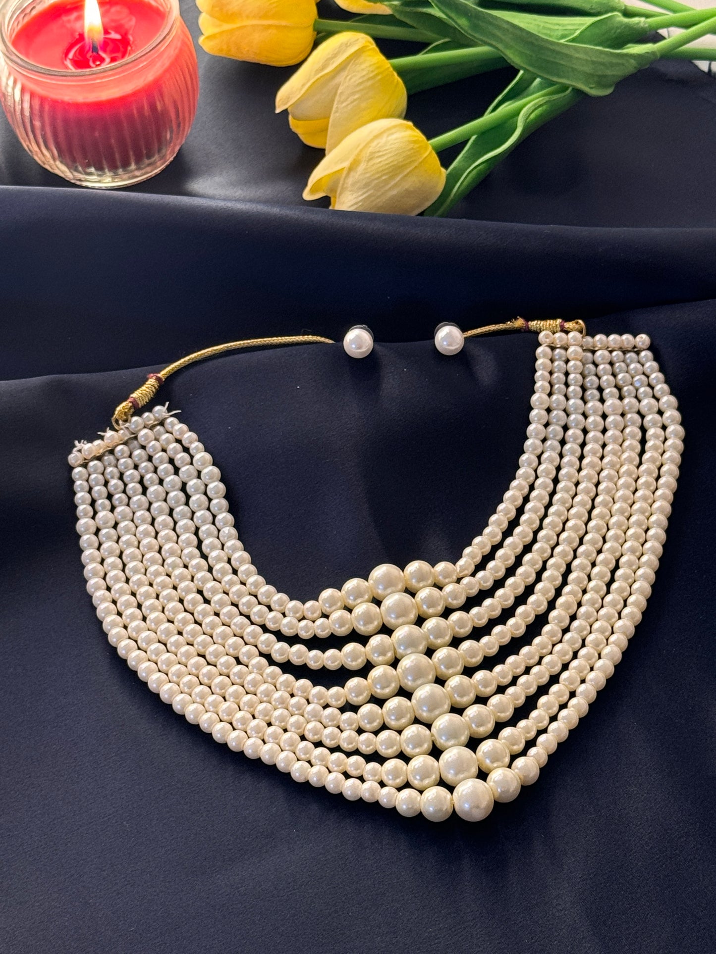 Pearl necklace on a dark fabric background with yellow tulips and a red candle.