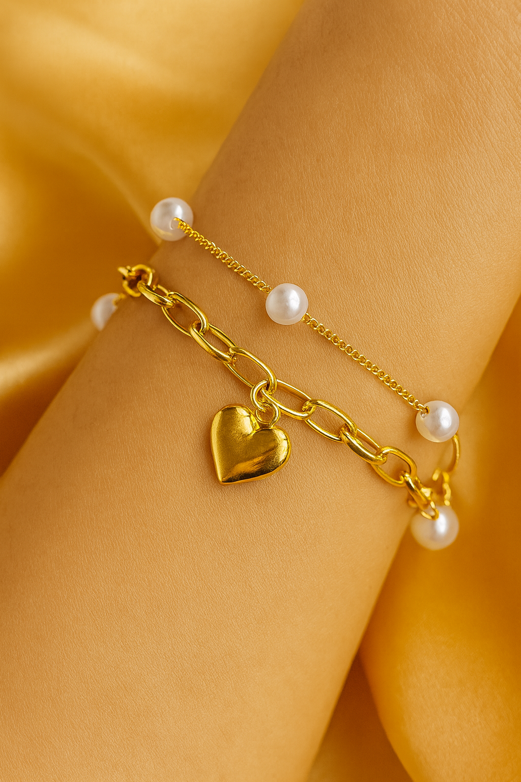 Gold bracelet with heart charm and pearls on a wrist against a yellow satin background