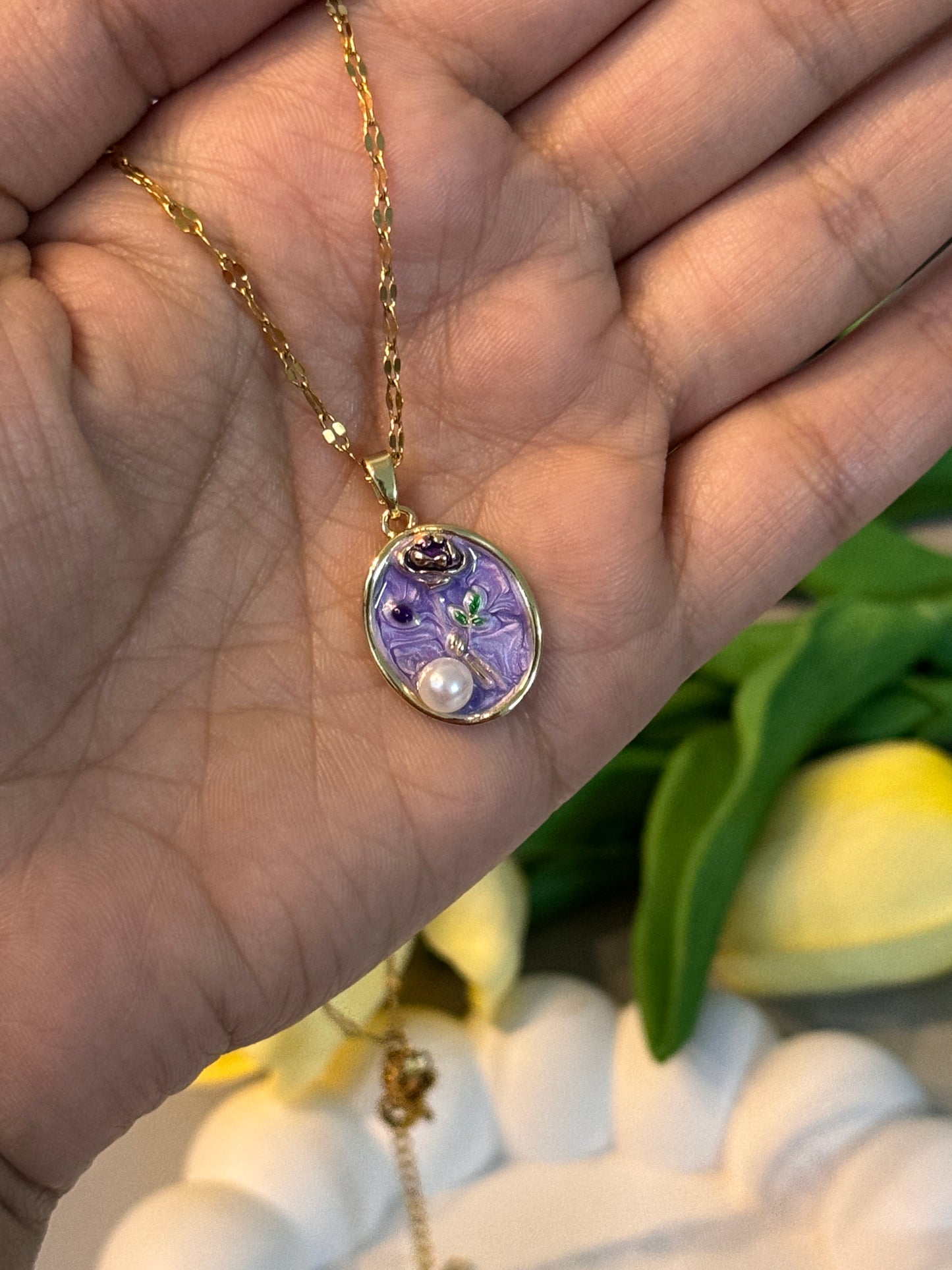 Gold necklace with a purple pendant held in a hand, with flowers in the background