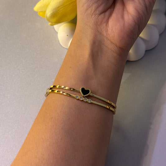 Hand wearing a gold bracelet with a heart charm, holding yellow flowers on a light background