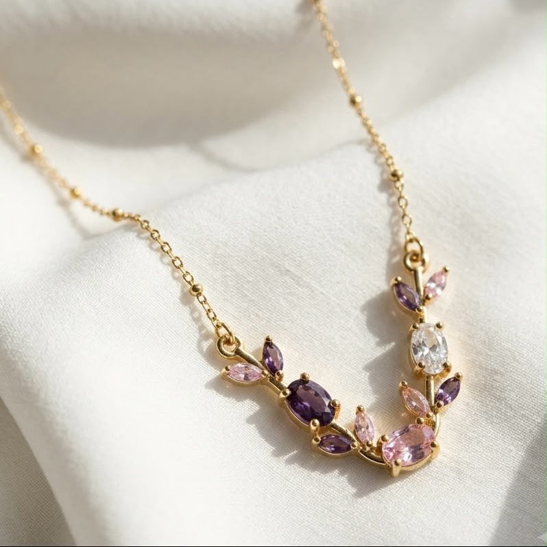 A gold leaf-shaped necklace with marquise-cut pink and purple gemstones, displayed on a soft white fabric background.