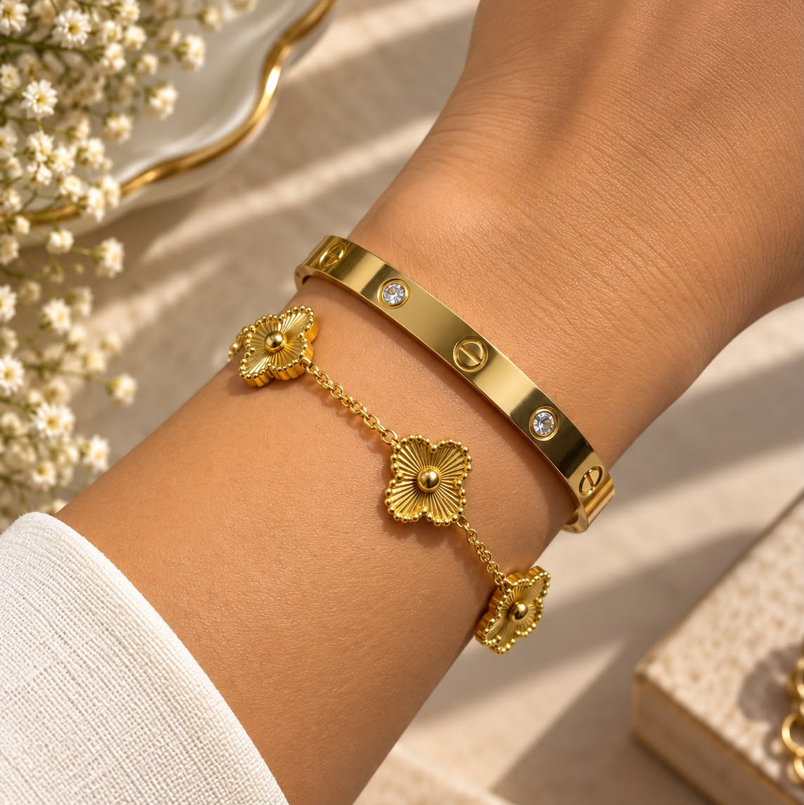 A close-up of a person's wrist wearing two gold bracelets: a slim Cartier-style bracelet with round bezel-set clear stones and a delicate chain bracelet with three gold flower charms.