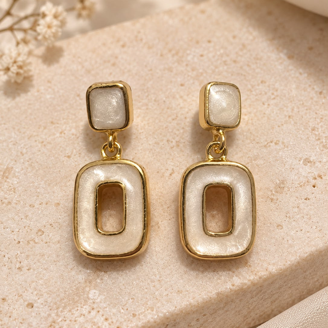 A pair of gold-tone drop earrings with rectangular mother-of-pearl inlays, displayed on a beige stone surface with dried flowers in the background.