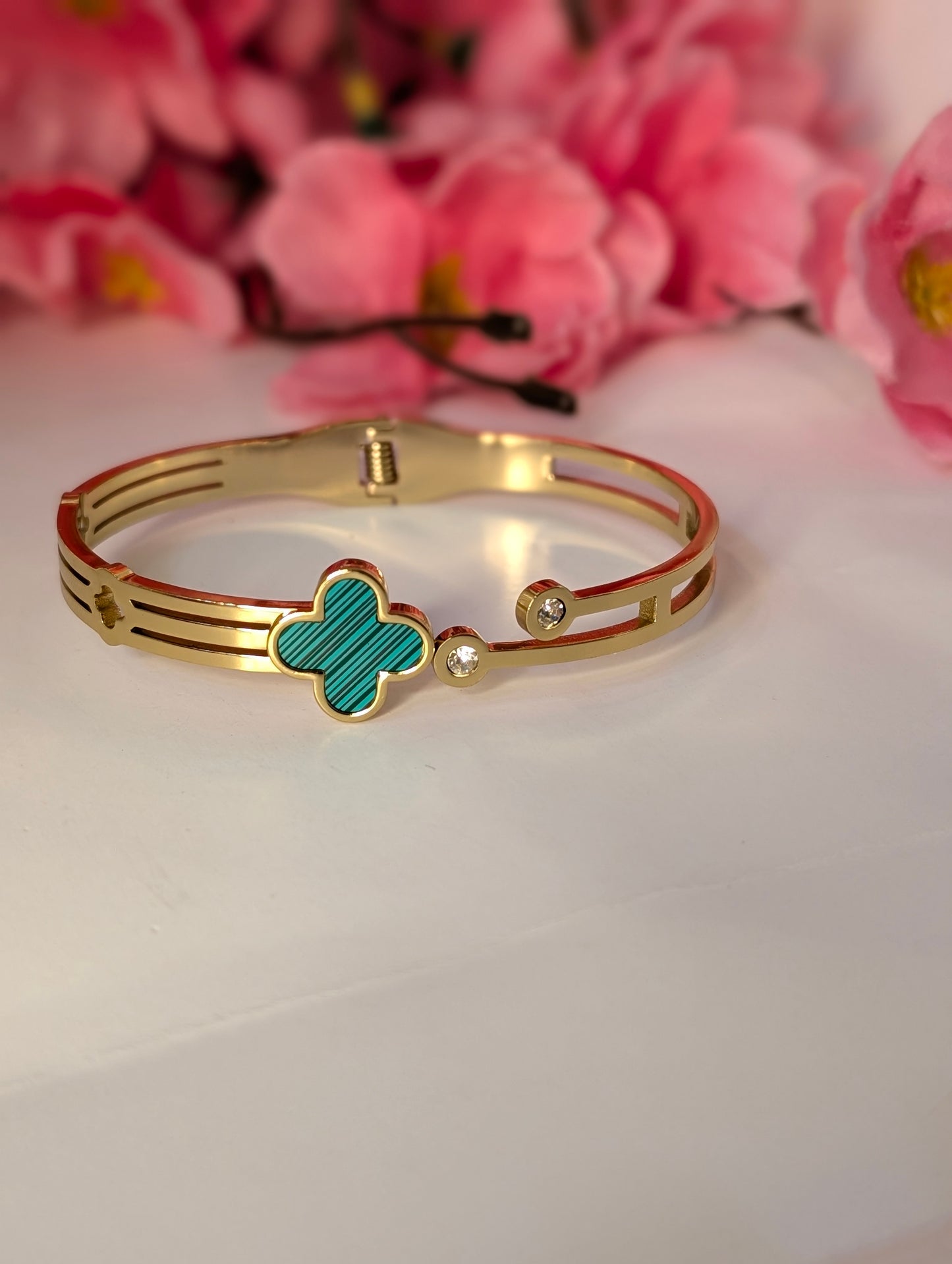 Gold bracelet with a green clover-shaped charm on a white surface with pink flowers in the background