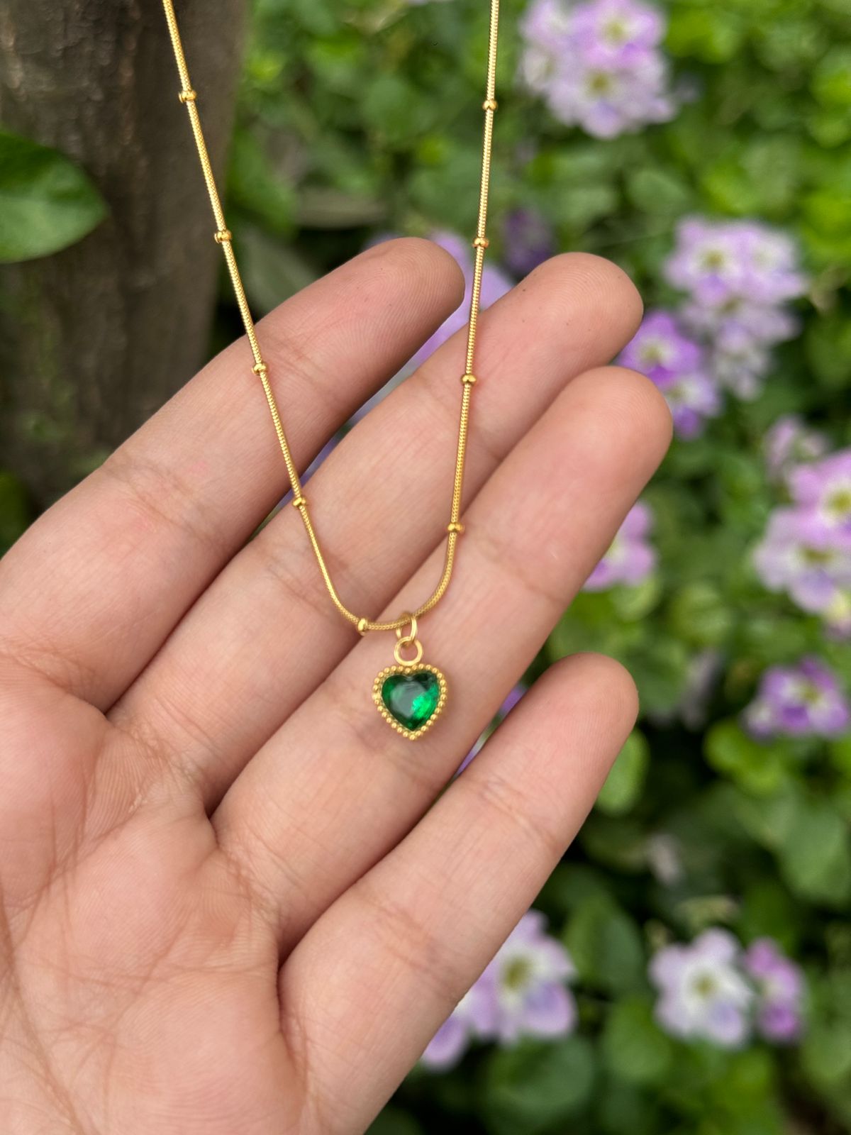 Gold necklace with a green heart pendant held in a hand against a blurred floral background