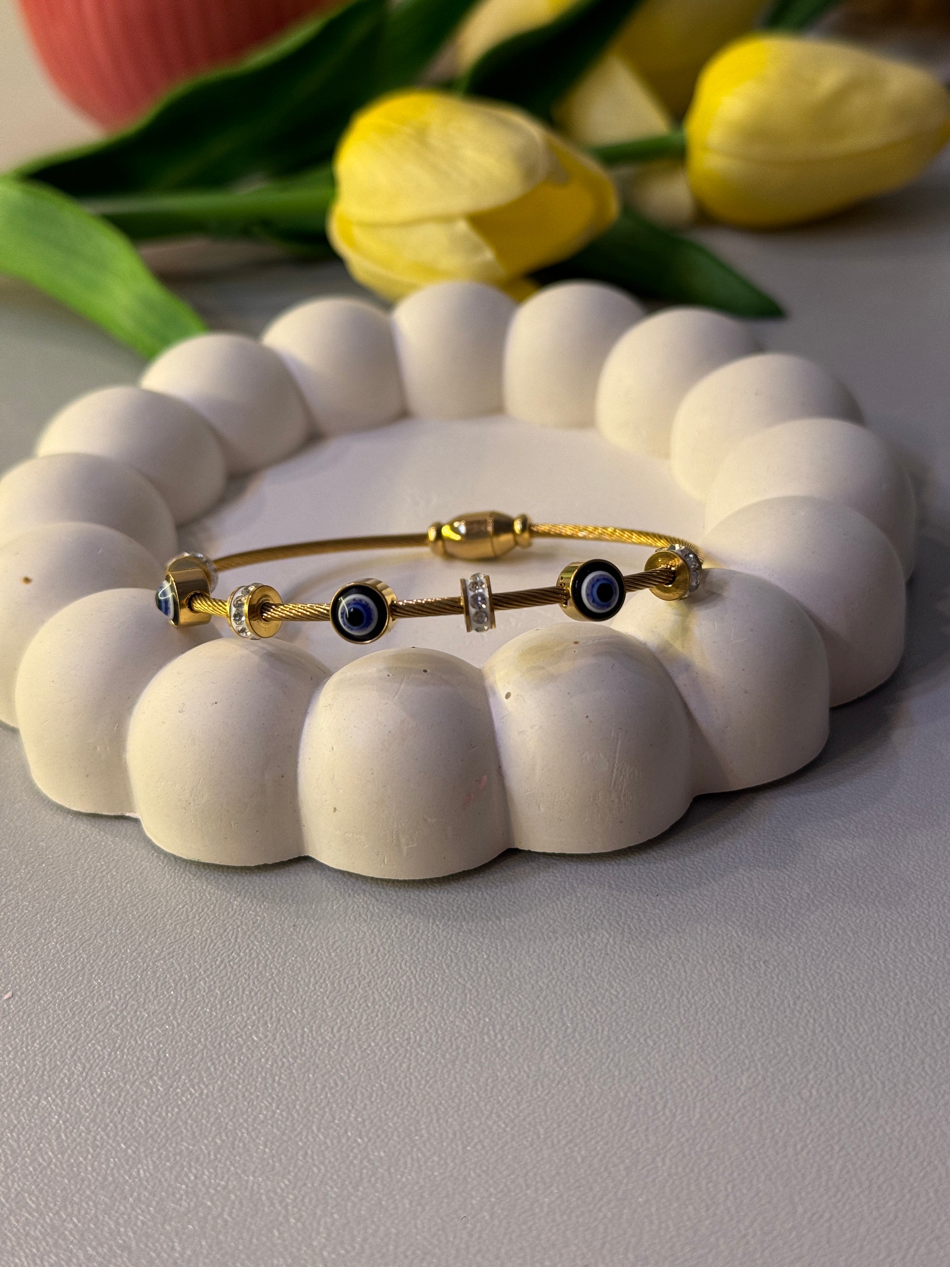 Gold bracelet with blue stones on a white beaded holder against a light gray background with yellow flowers.