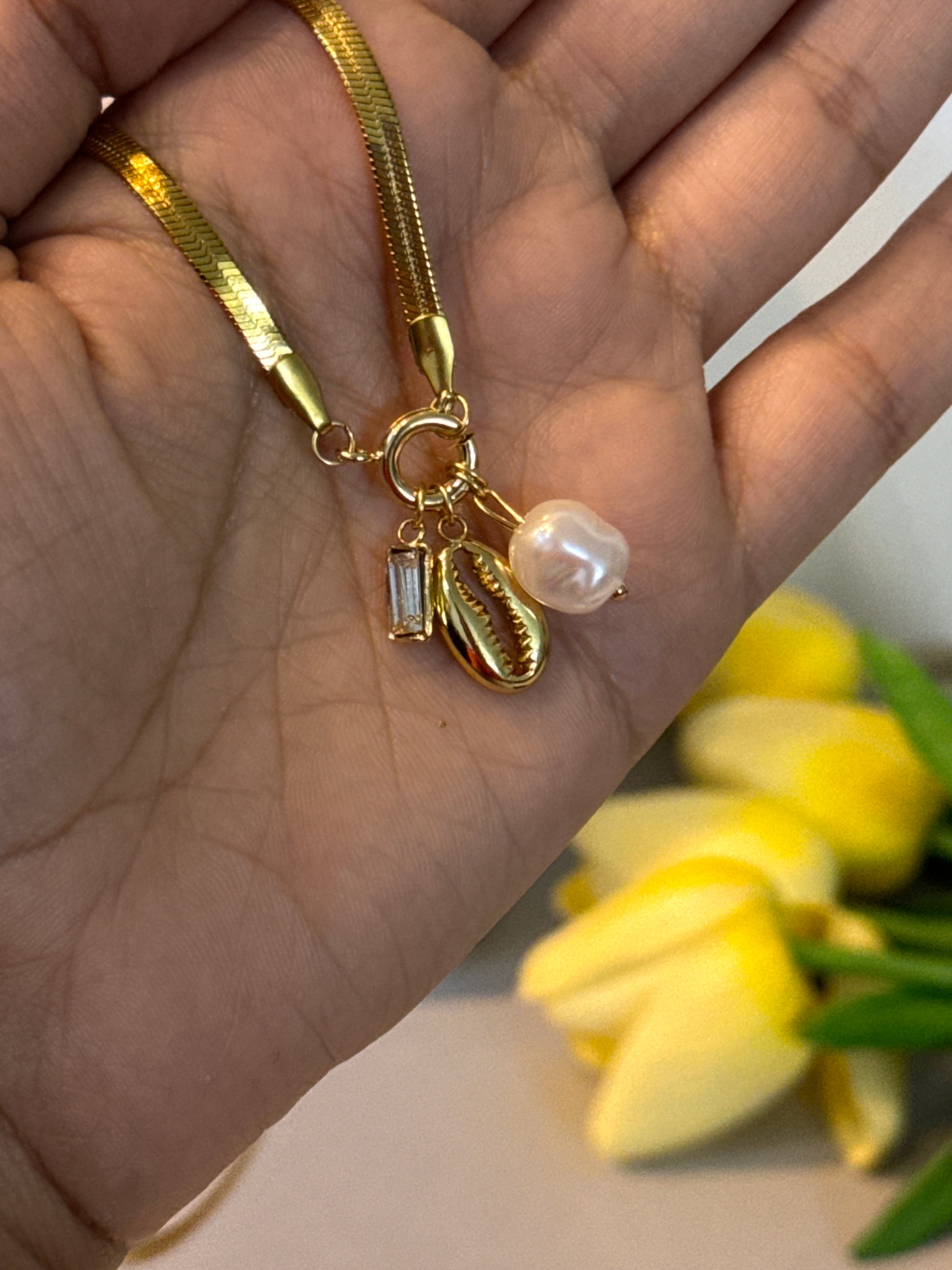 Gold bracelet with a pearl charm held in a hand, with yellow flowers in the background.